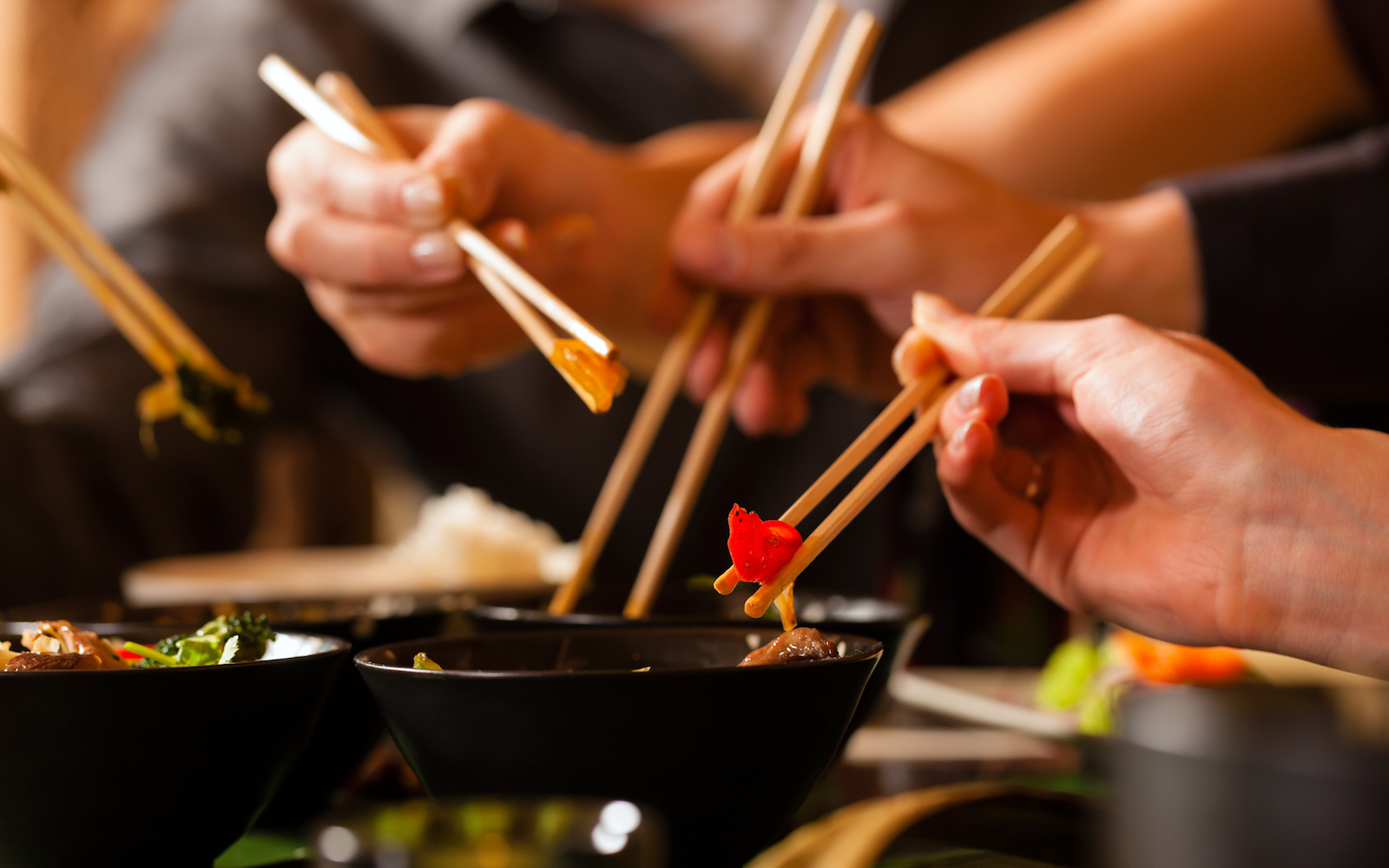 Four sets of chopsticks grabbing Asian food out of various black bowls sitting on a table in a Thai food restaurant.  Three pairs of chopsticks have food between them, and one is in a black bowl.  There is a person's chest out of focus in the background.  There are four sets of chopsticks visible, but there are only three hands visible.  There is a set of chopsticks coming into view from the left.  The hand in front is in focus, while the two hands in the background are slightly blurred.